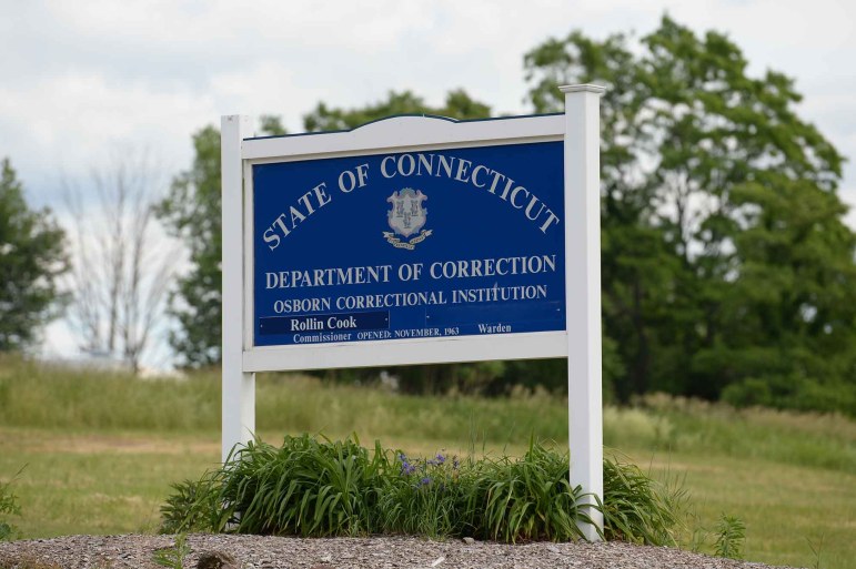 A blue sign in front of Osborn Correctional Institution. The blue sign sits on top of grass. Trees are in the background.