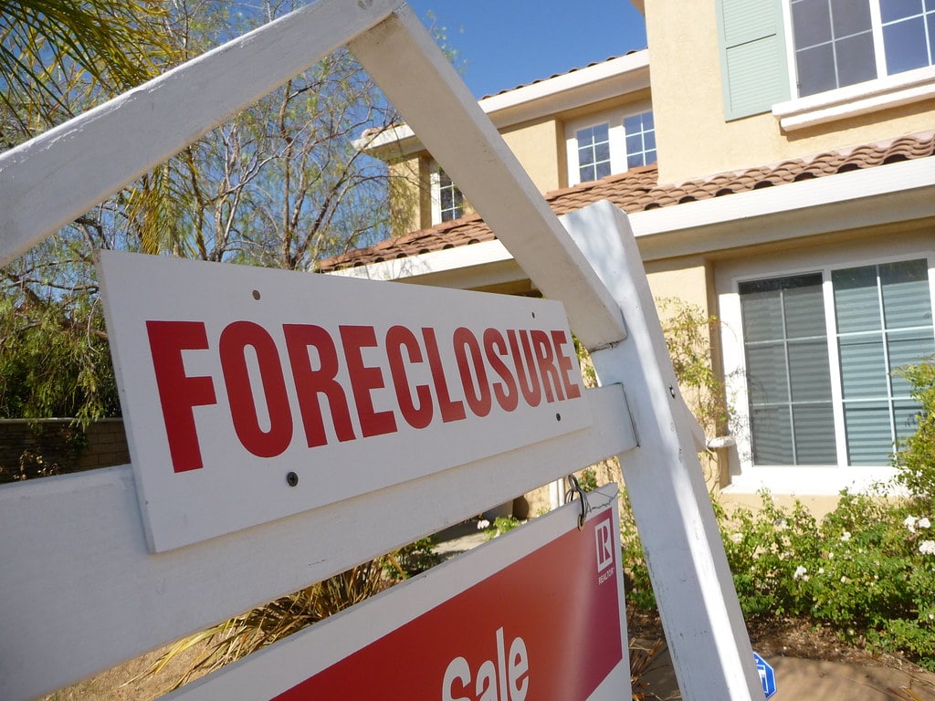 A foreclosure sign in the foreground in front of a cream-colored house.