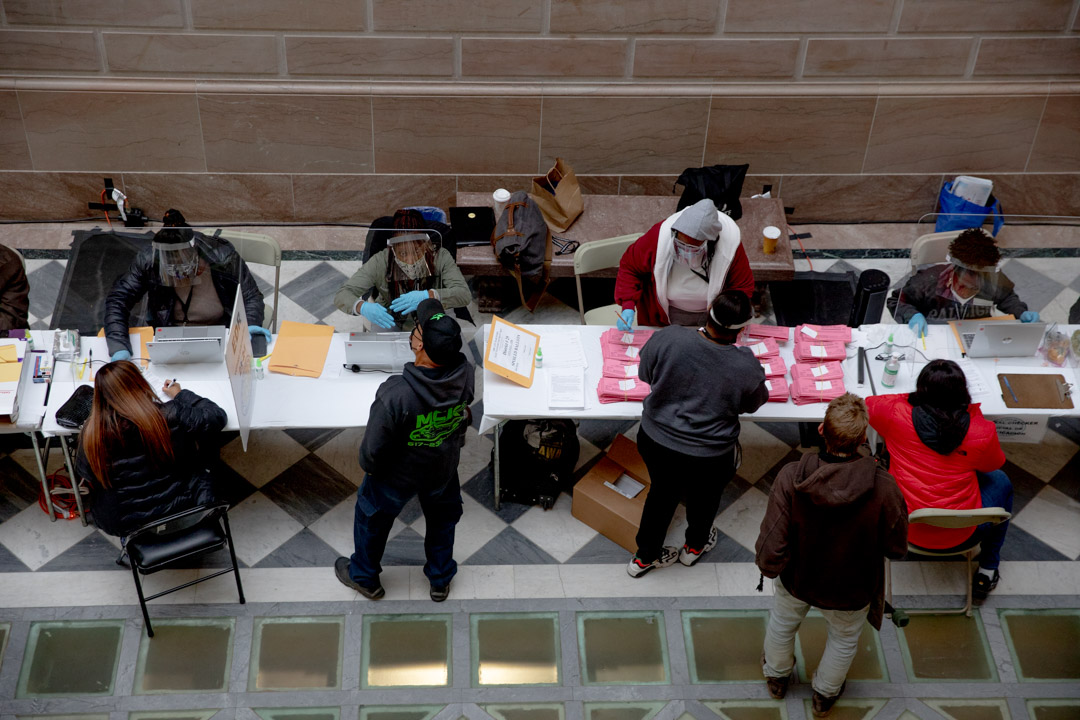 People are seen at a voting registration table interacting with election workers. Most people in the photos are wearing protective face masks.