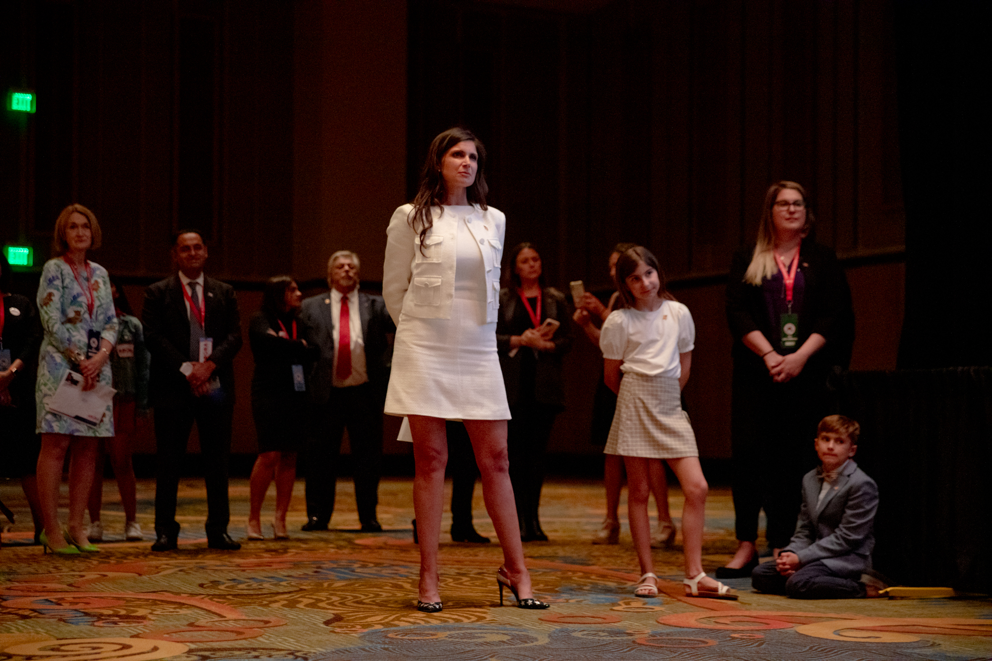 Jessica Kordas, wearing a white dress, stands in front of a group of several people, as she listens to someone else speak.