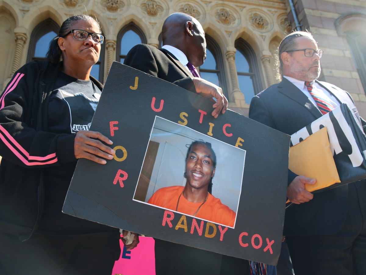 Doreen Coleman, the mother of Randy Cox, holds a Black sign with the words "Justice for Randy Cox," and a photo of her son smiling with a orange shirt and gold chain. Coleman is wearing glasses and a black jacket with pink stripes.
