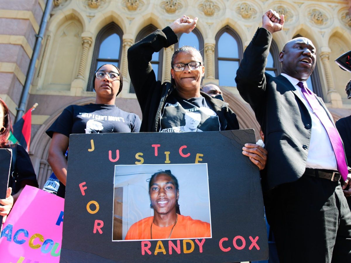 Doreen Coleman, the mother of Randy Cox, holds a sign that says "Justice for Randy Cox," stands with her fist raised in the air. To her right is civil rights attorney Ben Crump, who's also holding his fist up. It's a sunny day outside.