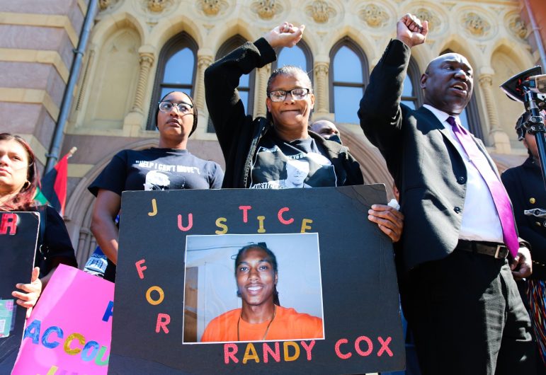 Doreen Coleman, the mother of Randy Cox, holds a sign that says "Justice for Randy Cox," stands with her fist raised in the air. To her right is civil rights attorney Ben Crump, who's also holding his fist up. It's a sunny day outside.