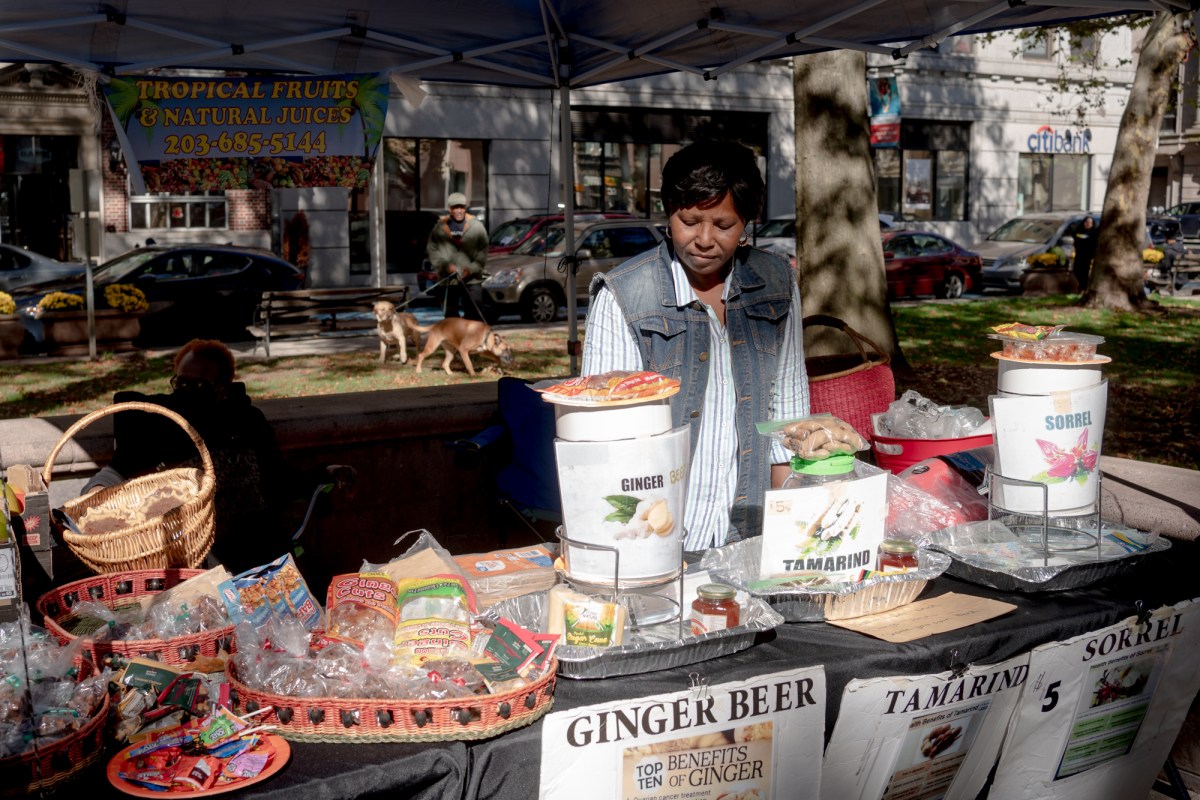 A woman working at a farmer's market hands stands under her tent, near tables topped with snacks and drinks.