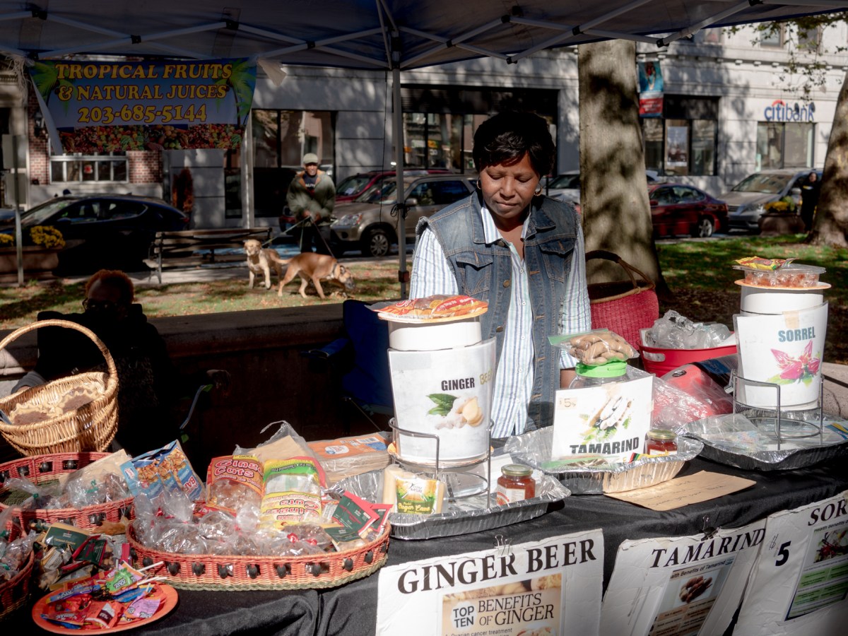 A woman working at a farmer's market hands stands under her tent, near tables topped with snacks and drinks.
