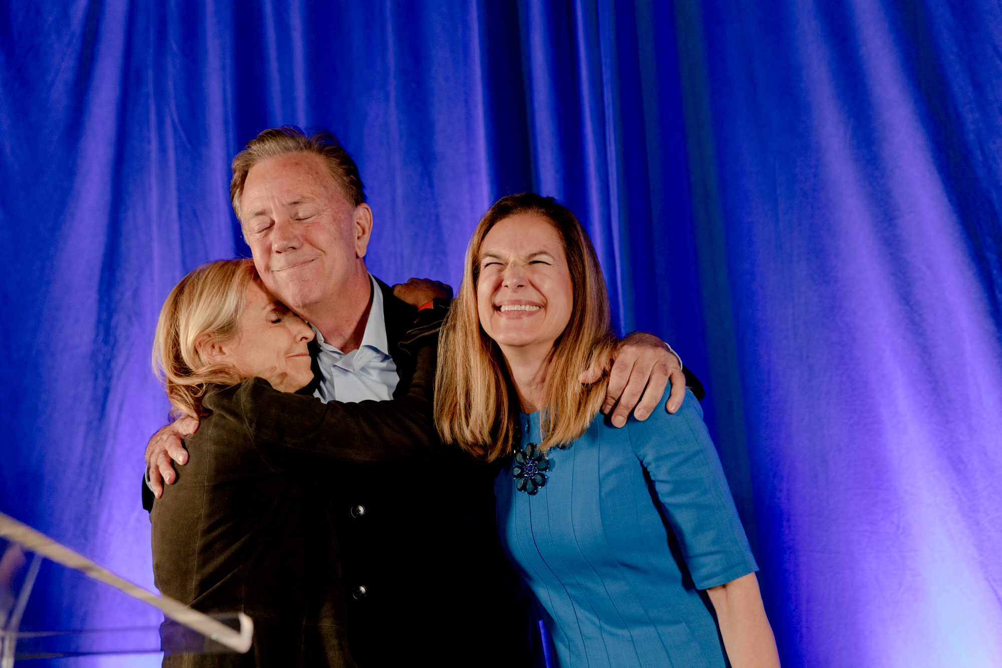 El gobernador Lamont se para contra un fondo azul con un brazo alrededor de su esposa y el otro alrededor de la vicegobernadora.