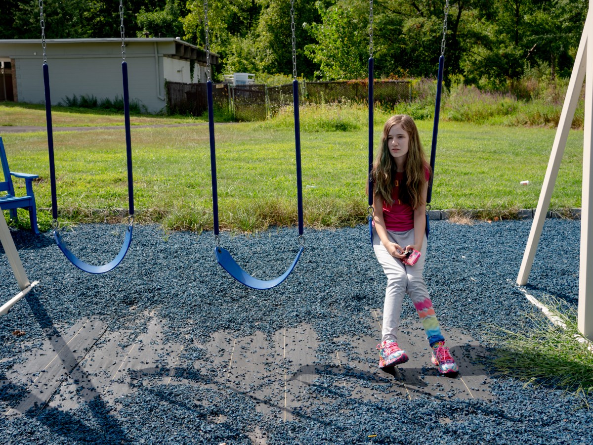 A girl sits alone on a swing set. It's a sunny day and grass is behind her.