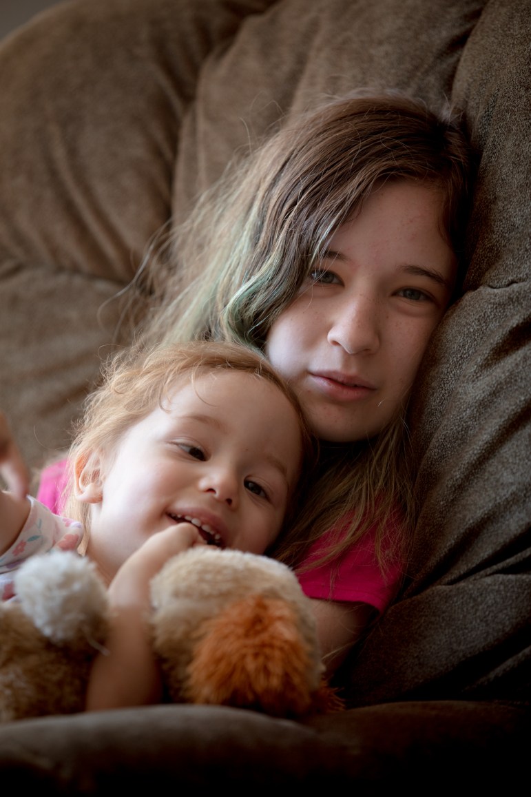 A teenage girl holds her 2-year-old sister on a couch. The toddler is holding a stuffed animal