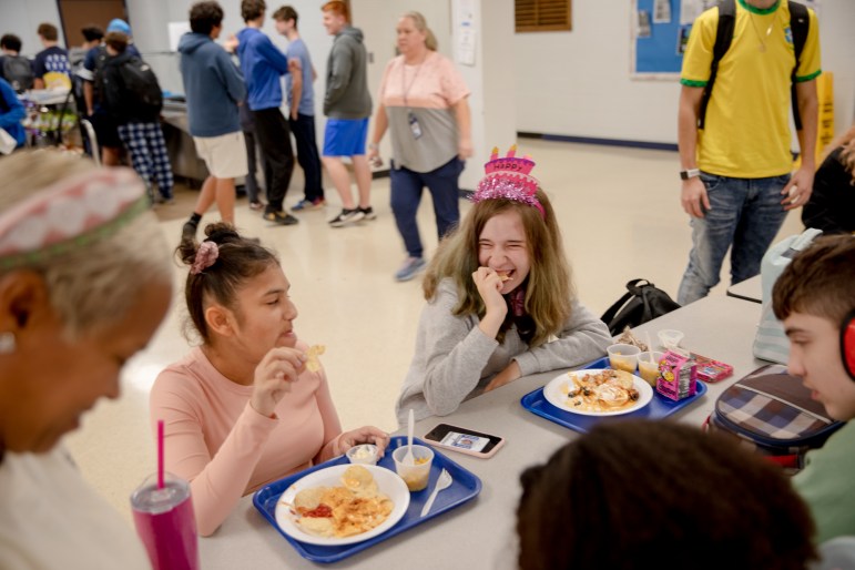 Two teenage girls sit and eat lunch together at a school cafeteria. One is wearing a pink birthday hat shaped like a cake and laughing.