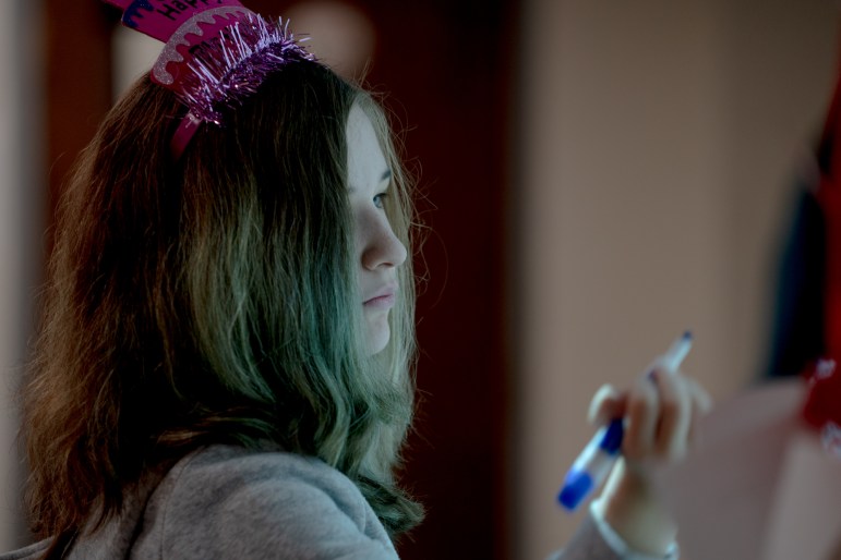 A teenage girl in a pink birthday hat holds a blue dry erase marker, getting ready to write on a board.