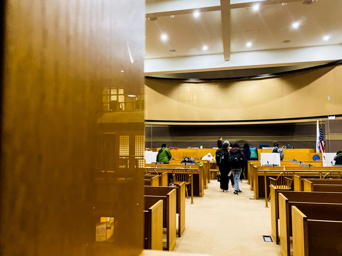 A group of people wait in a short line inside of New Haven City Hall. The lights are on. Private voting stations, which look like large privacy folders, are seen in the distance.