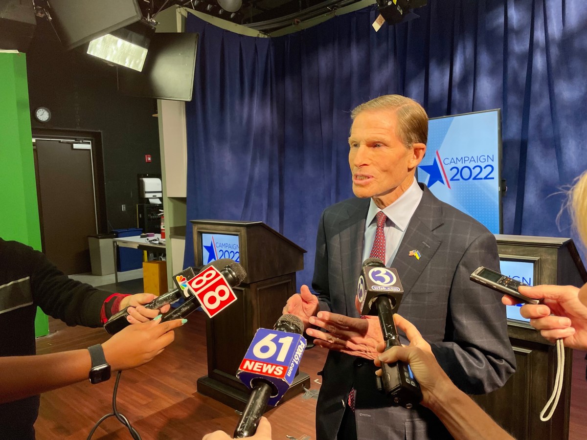 Several microphones are held in front of U.S. Senator Richard Blumenthal, who is standing in front of a podium following a debate in the 2022 U.S. Senate race.