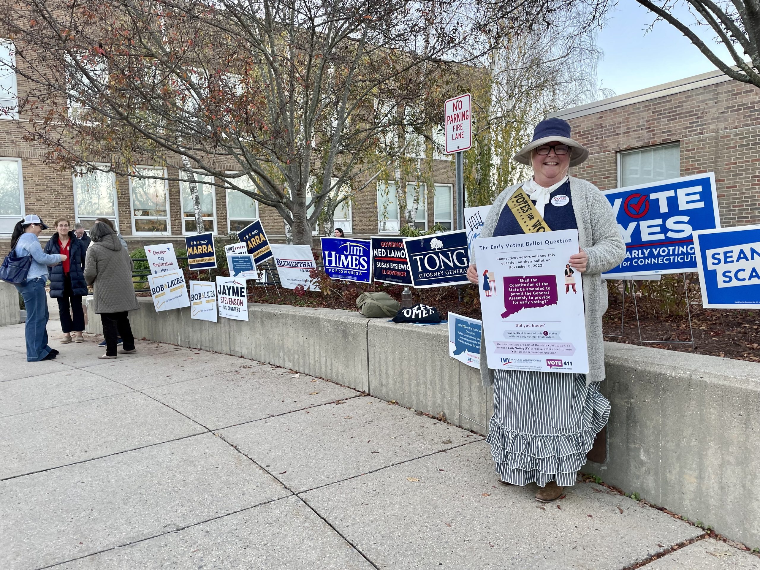 A woman dressed as a suffragist stands in front of a polling location at Darien Town Hall. Campaign signs line the wall behind her.