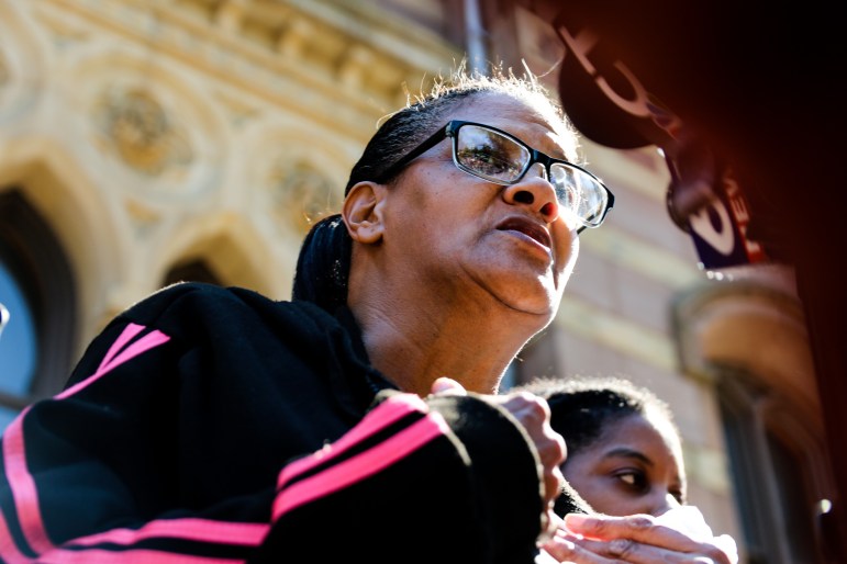 Doreen Coleman, a woman wearing glasses and a black jacket with pink stripes, speaks in front of a microphone.