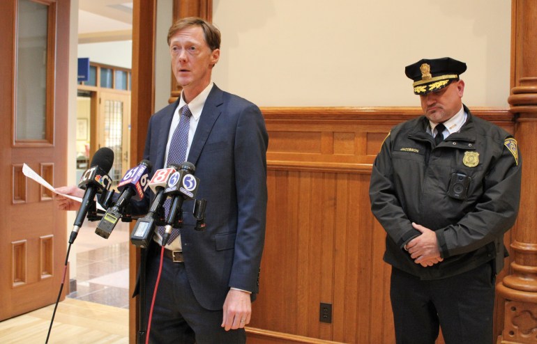 New Haven Mayor Justin Elicker stands in front of TV news microphones while holding a piece of paper and speaking with the media, while police chief Karl Jacobson stands nearby listening with one hand crossed over the other.