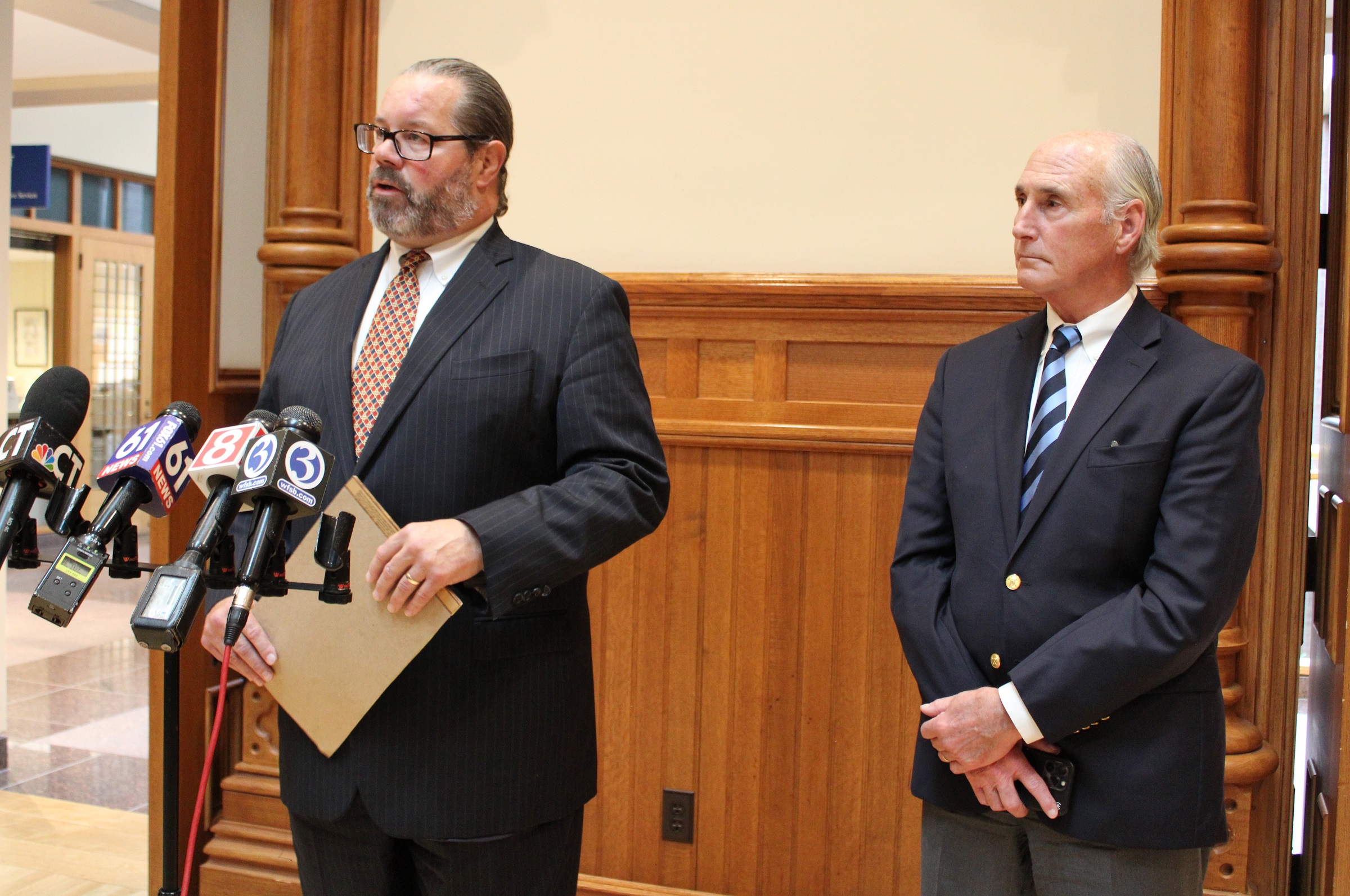 Randy Cox's attorney, standing next to his colleague, stands in front of TV news microphones while holding a notepad, answering questions from members of the media.