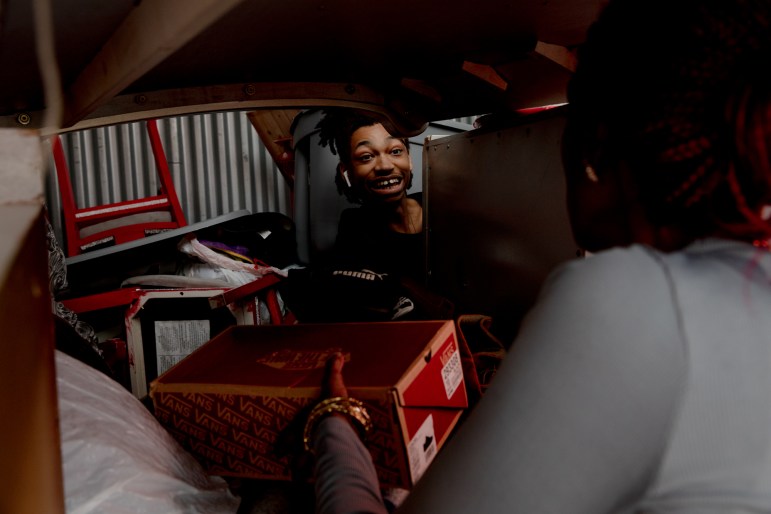 A teenage boy looks into a mirror in a crowded storage unit. He is smiling.