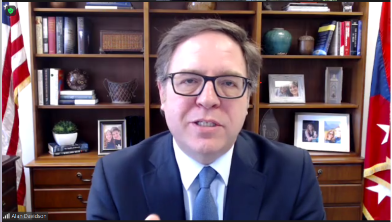 Assistant Secretary of Commerce Alan Davidson speaks at his desk with a bookshelf and flags in the background.