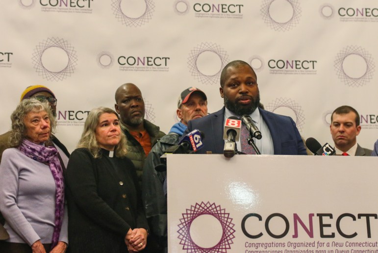 Sen. Gary Winfield, in a suit and tie, stands in front of a podium with microphones to speak, while several people stand behind him to watch along.