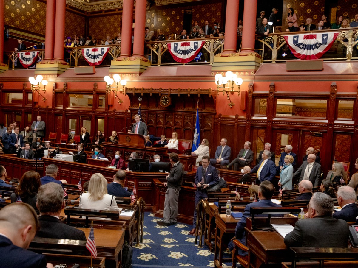 Connecticut state representatives are gathered on the House floor of the State Capitol Building. House Speaker Matt Ritter is seen at the front of the room, behind a podium, addressing his colleagues.