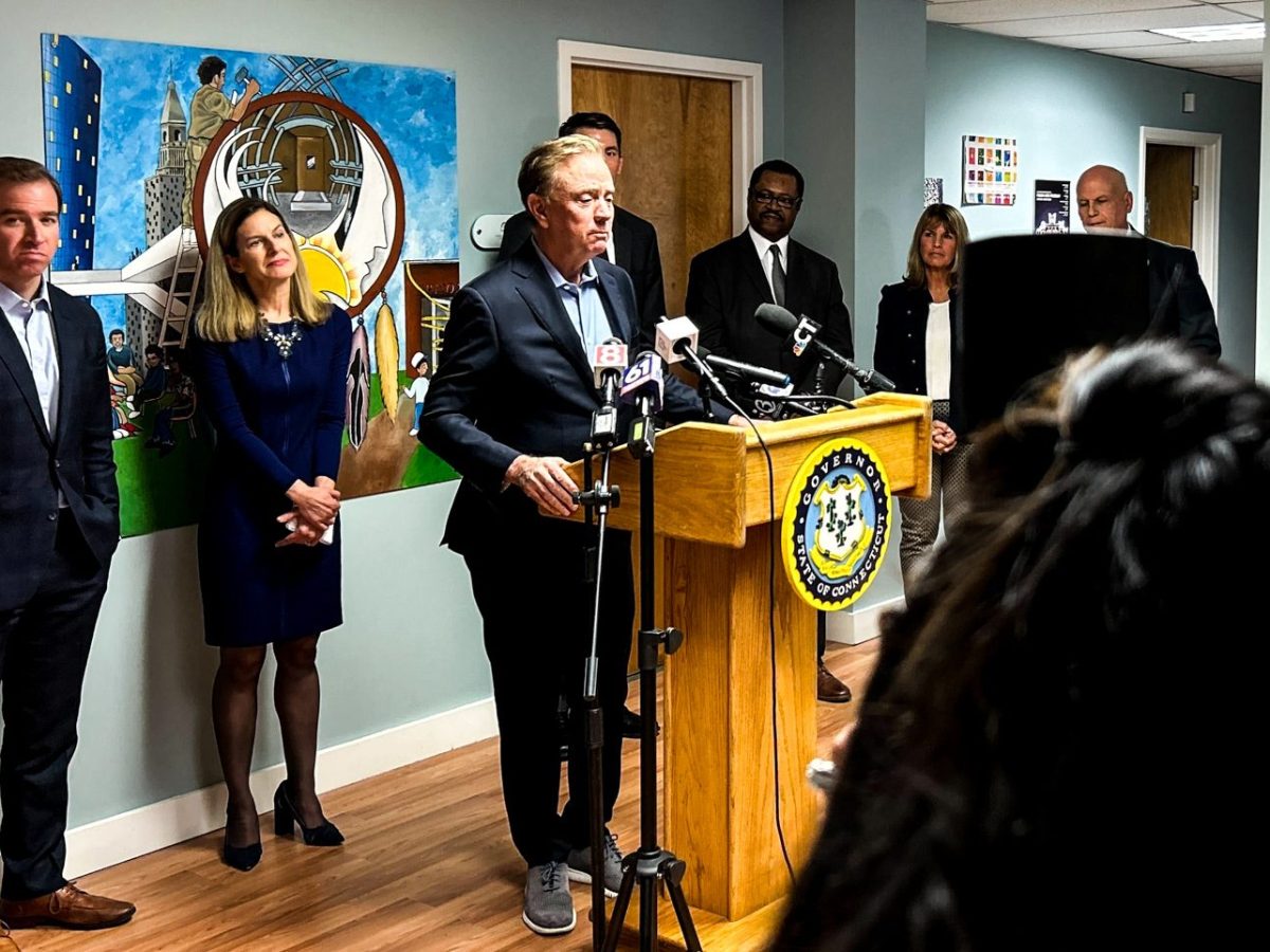 Gov. Ned Lamont, dressed in formal attire, stands in front a podium with microphones — with several city and state officials standing behind him. He's speaking in a room full of reporters and advocates.