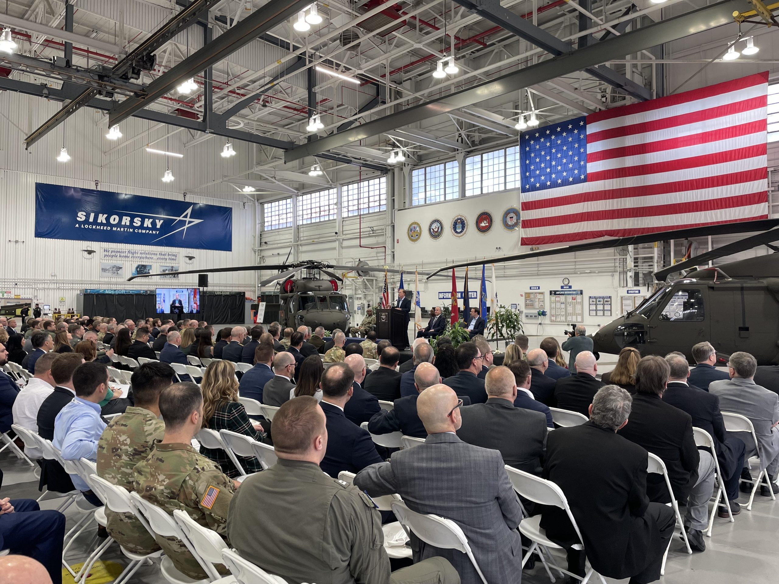 A large gathering of people in an airplane hangar, with two helicopters on display and a man at a podium speaking.