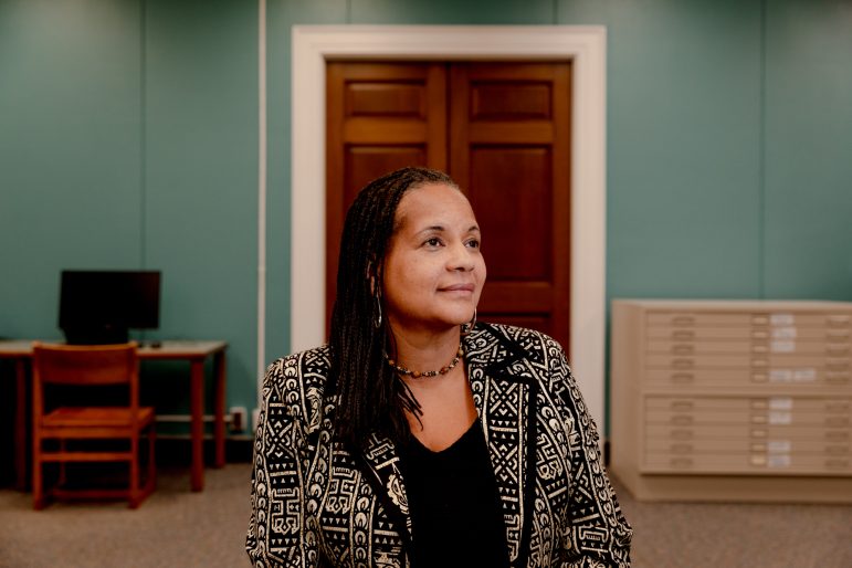 Una mujer, sentada, con un blazer blanco y negro. Detrás de ella hay una puerta y un escritorio con una computadora.