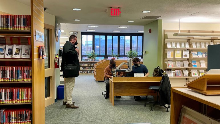 Tres personas entre los estantes de una biblioteca pública. Dos están sentados en un escritorio. Uno está de pie.