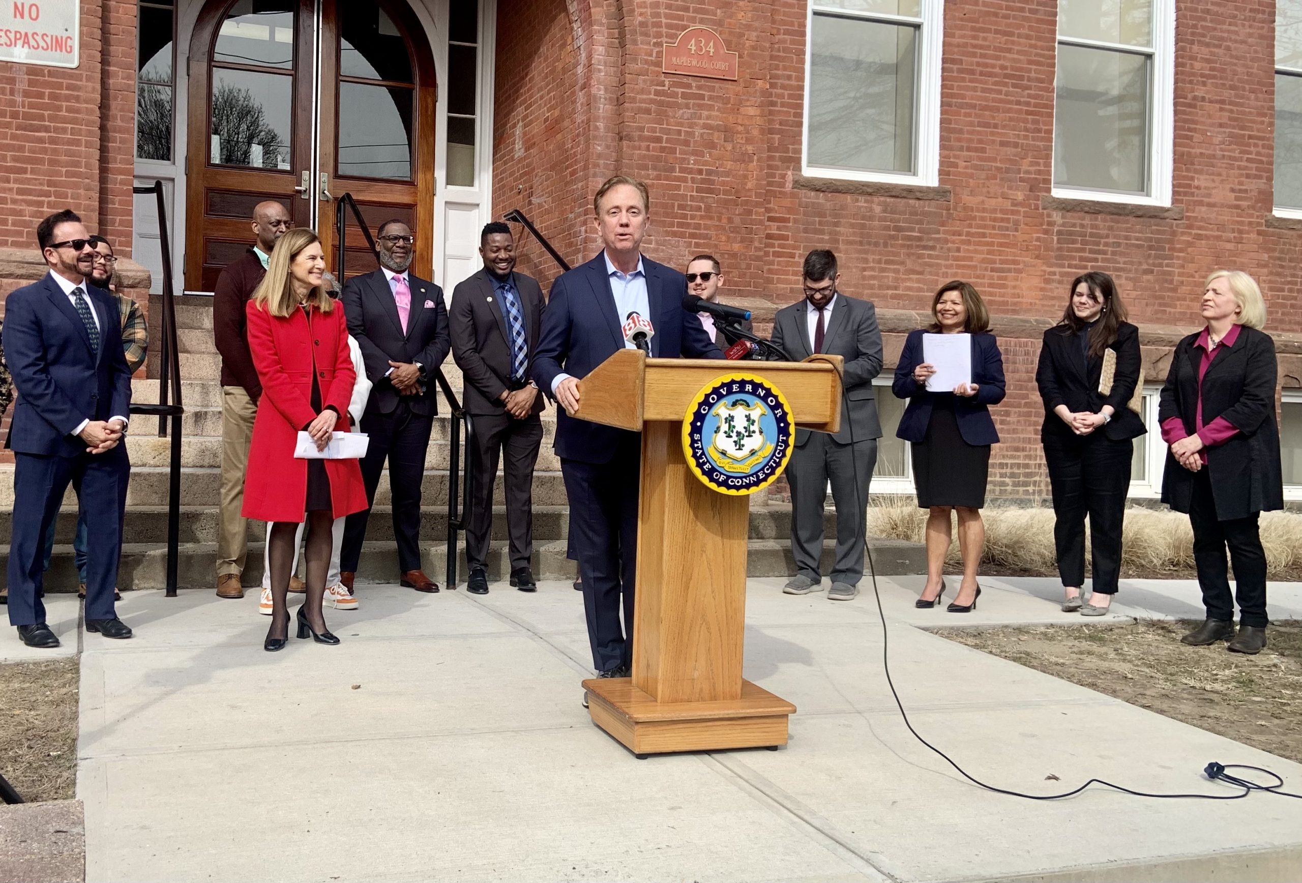 Gov. Ned Lamont stands in front of a building in Bridgeport with other officials on either side.