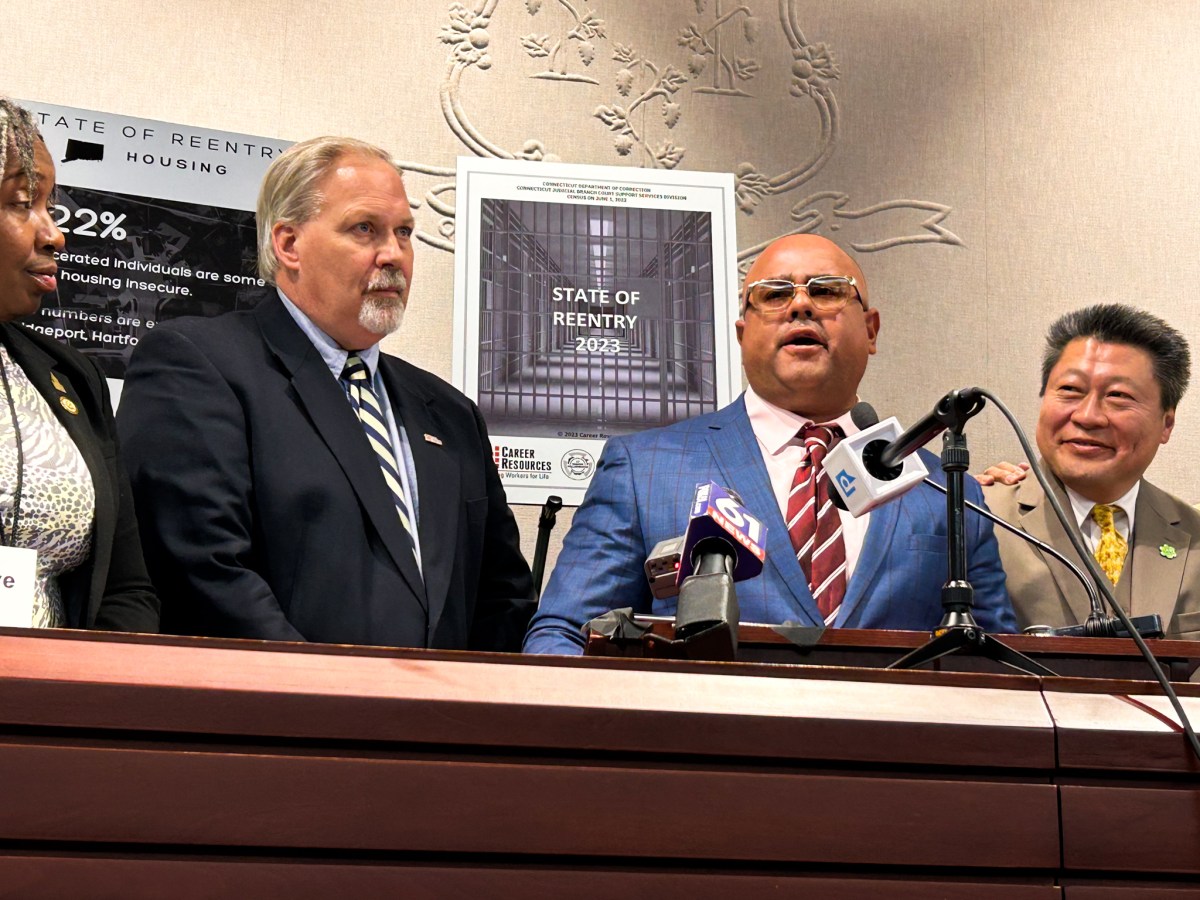 Virgilio Rosario, wearing a navy blue suit jacket and tinted eyeglasses, stands in front of a podium full of microphones and speaks to a room of people.