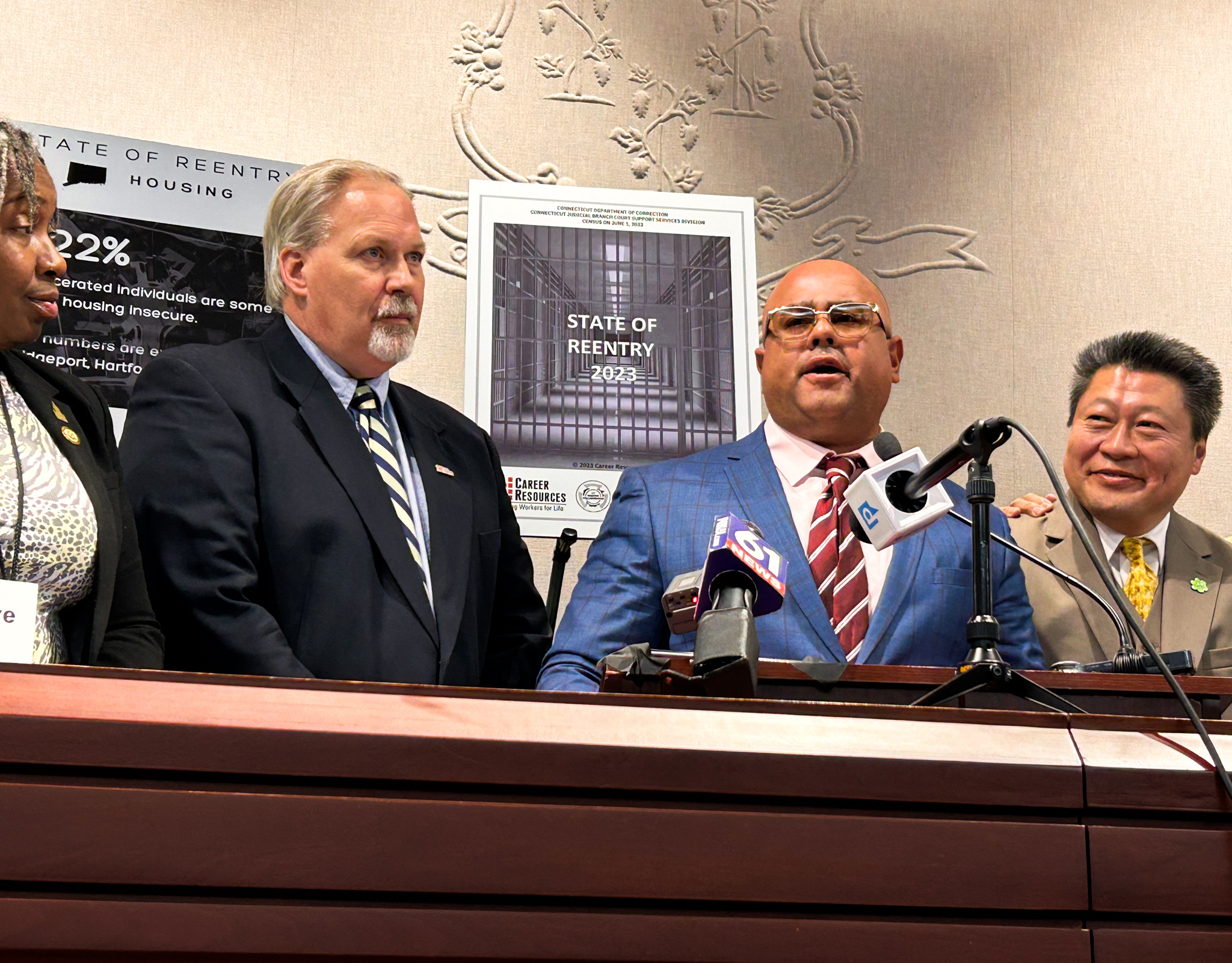 Virgilio Rosario, wearing a navy blue suit jacket and tinted eyeglasses, stands in front of a podium full of microphones and speaks to a room of people.