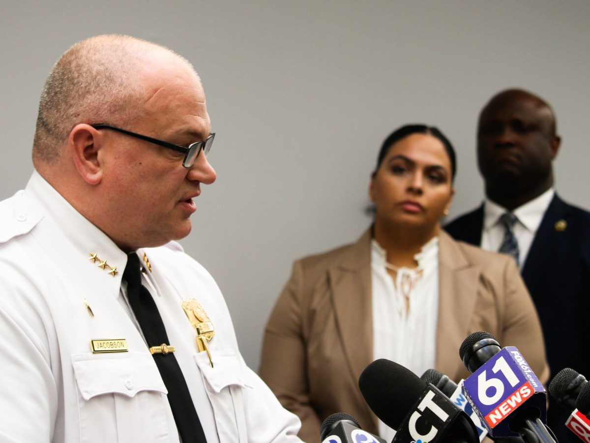 New Haven Police Chief Karl Jacobson, wearing a white uniform with a black tie, is seen standing a podium with two of his colleagues watching from behind.