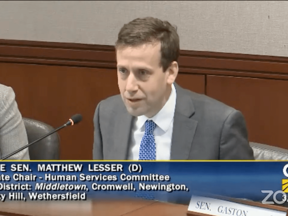 A man dressed in a suit sits at a table while speaking into a microphone.