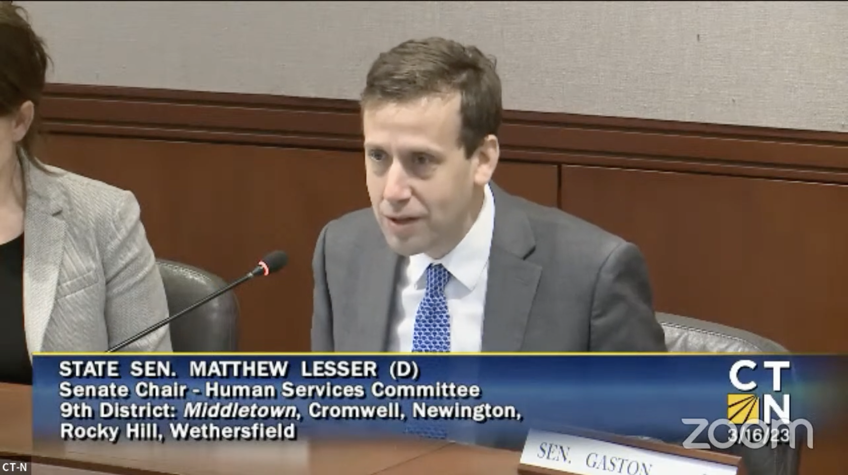 A man dressed in a suit sits at a table while speaking into a microphone.