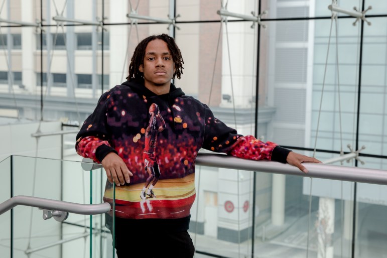Arjae Hill, a student at UConn Stamford, stands against a railing while posing for a photo on campus. He's wearing a sweater that shows Michael Jordan attempting a shot.