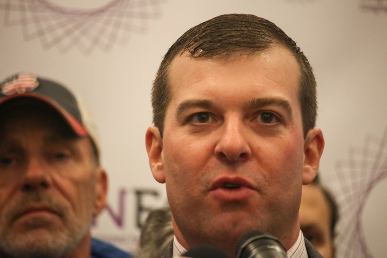 Rep. Steven Stafstrom looks ahead as he speaks into a microphone. Two people are seen standing behind him.