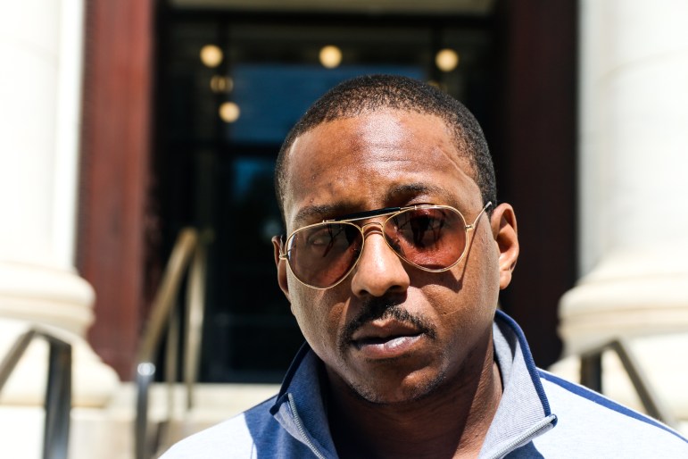 Jewu Richardson, wearing sun glasses and a blue-striped gray polo sweater, poses for a portrait photo in front of a New Haven courthouse.