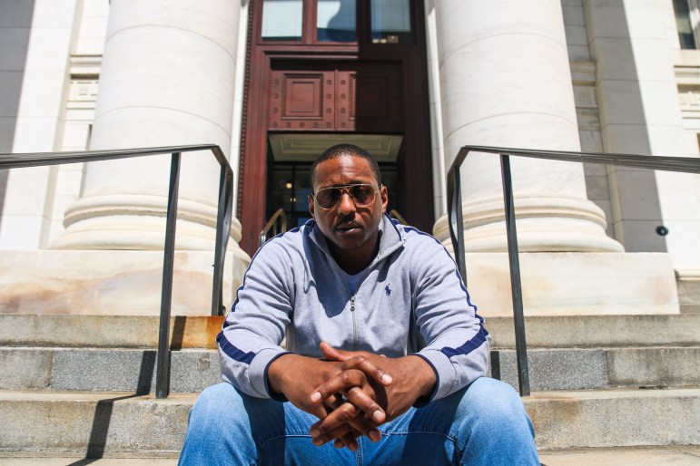 Jewu Richardson, wearing sun glasses and a blue-striped gray polo sweater, sits on the steps of a New Haven courthouse.
