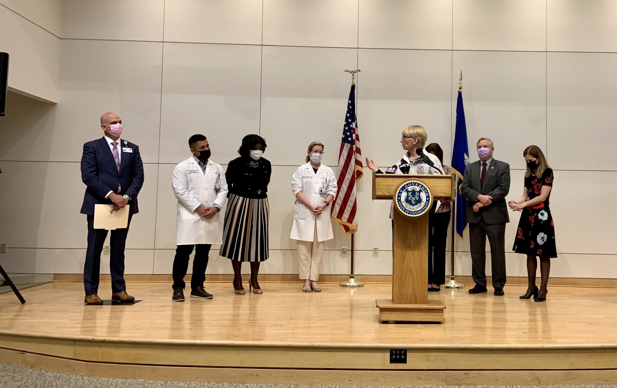 A woman speaks from a podium on a stage with other people standing on either side of her.