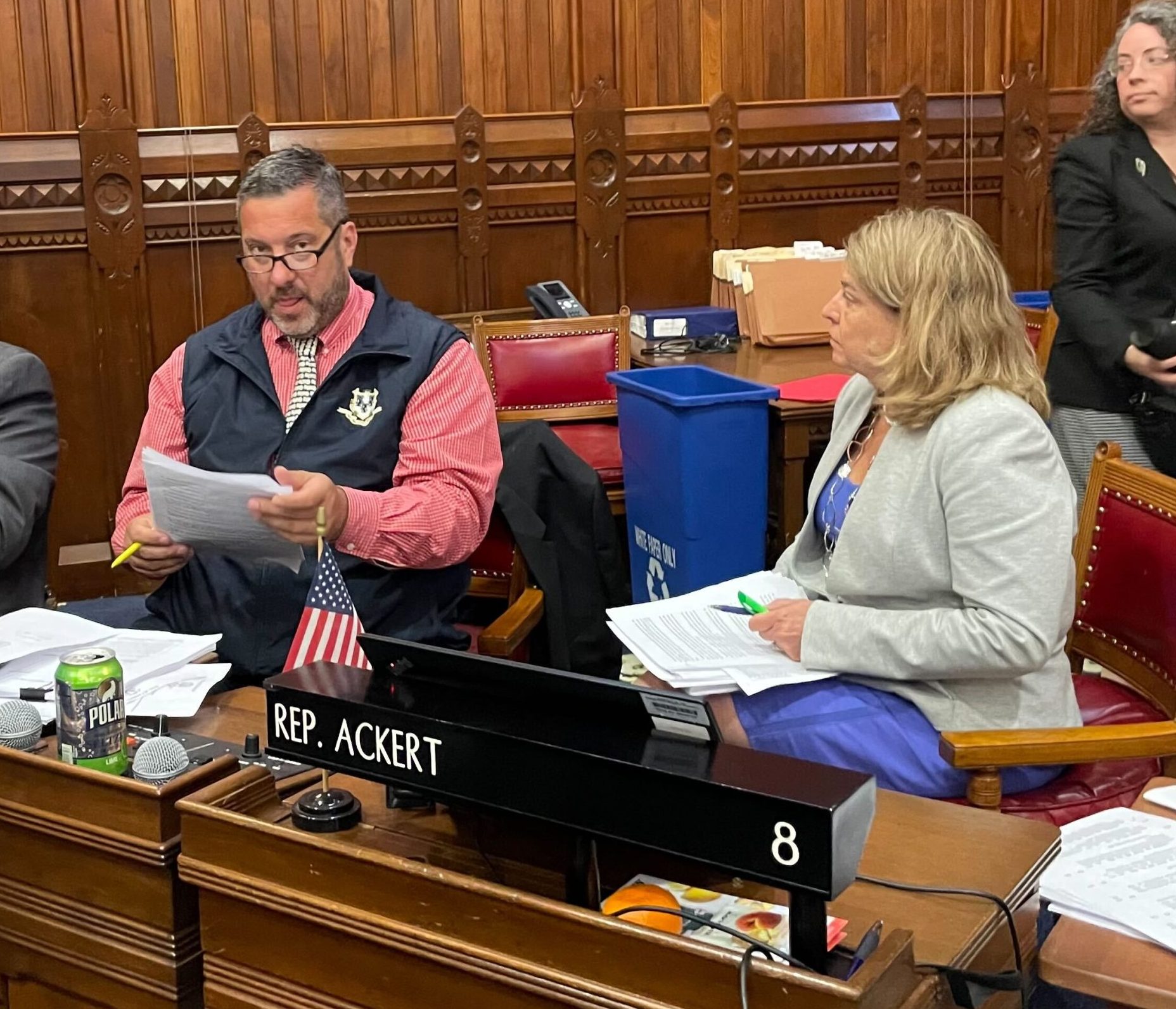 Two people sit at a desk in a wood paneled room.