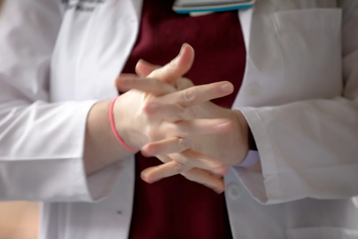 Las manos de una persona juntas con los dedos extendidos. La persona lleva una camisa roja y una bata de laboratorio blanca.