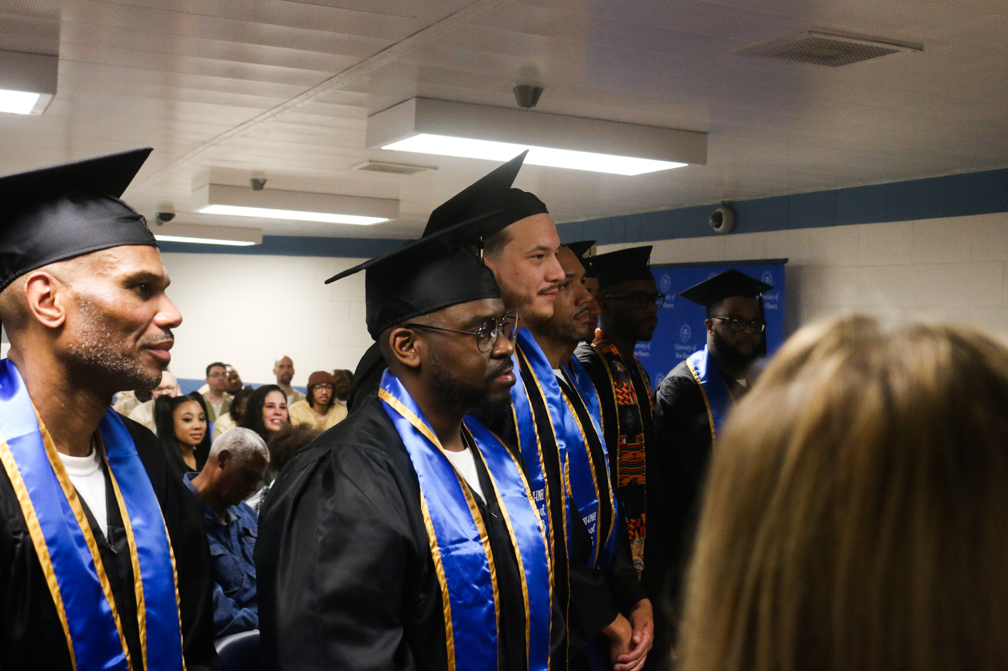 Seven people who've been incarcerated at MacDougall-Walker Correctional Institution, wearing black cap and gowns, listen to their commencement speakers in a dimly-lit room.