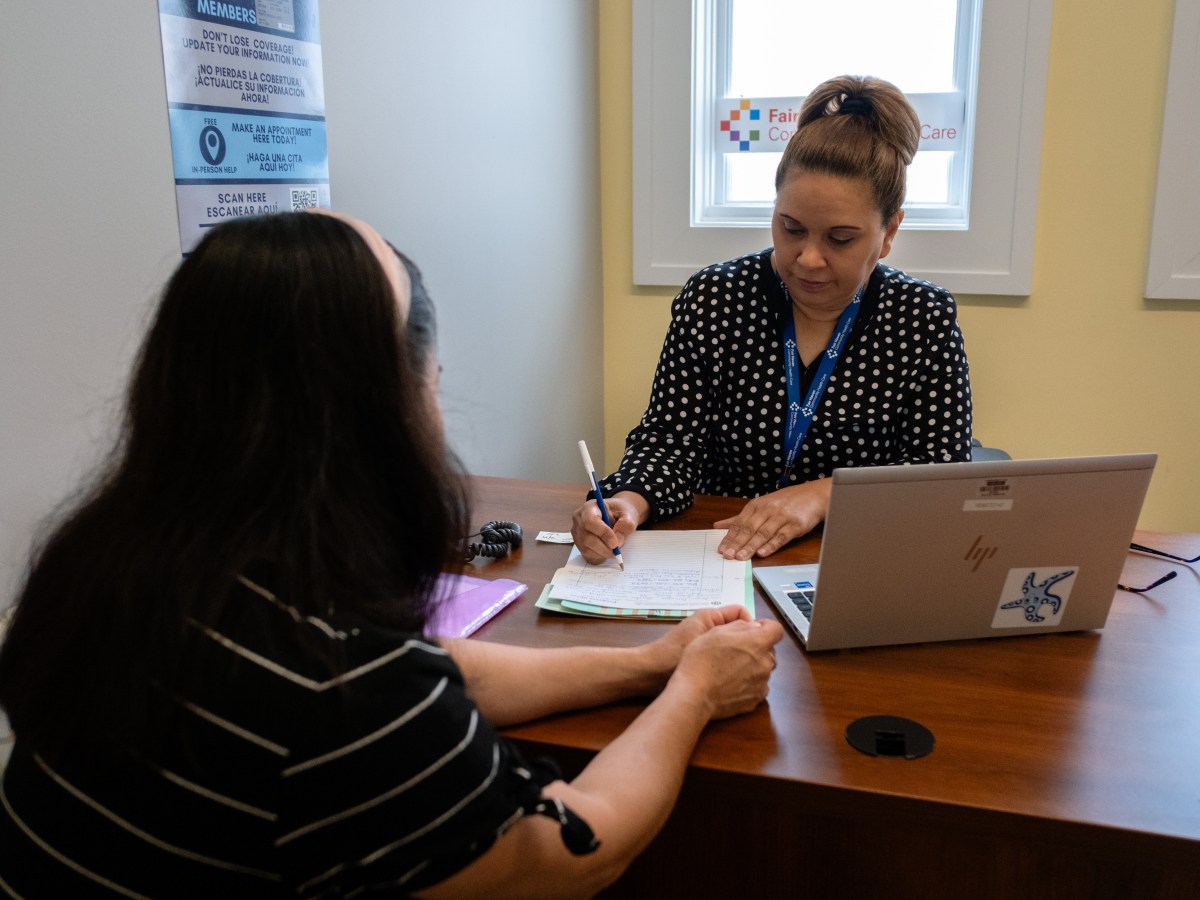 Two people sit across a desk from one another. A laptop sits open on the desk.