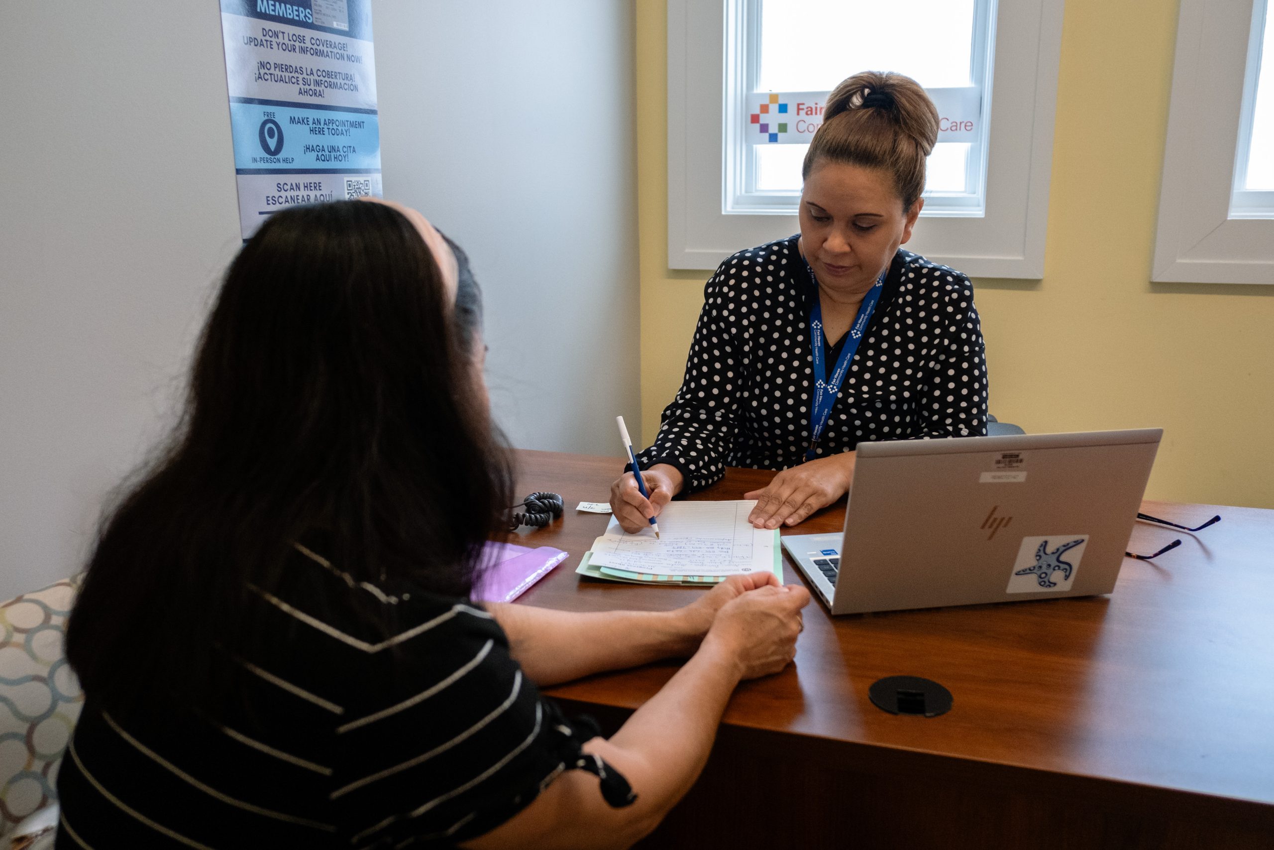 Two people sit across a desk from one another. A laptop sits open on the desk.