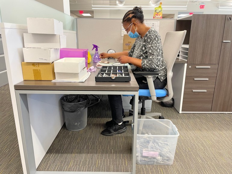 A woman seated at a desk with a spray bottle, cleaning cloths and a tray of designer eyeglasses.