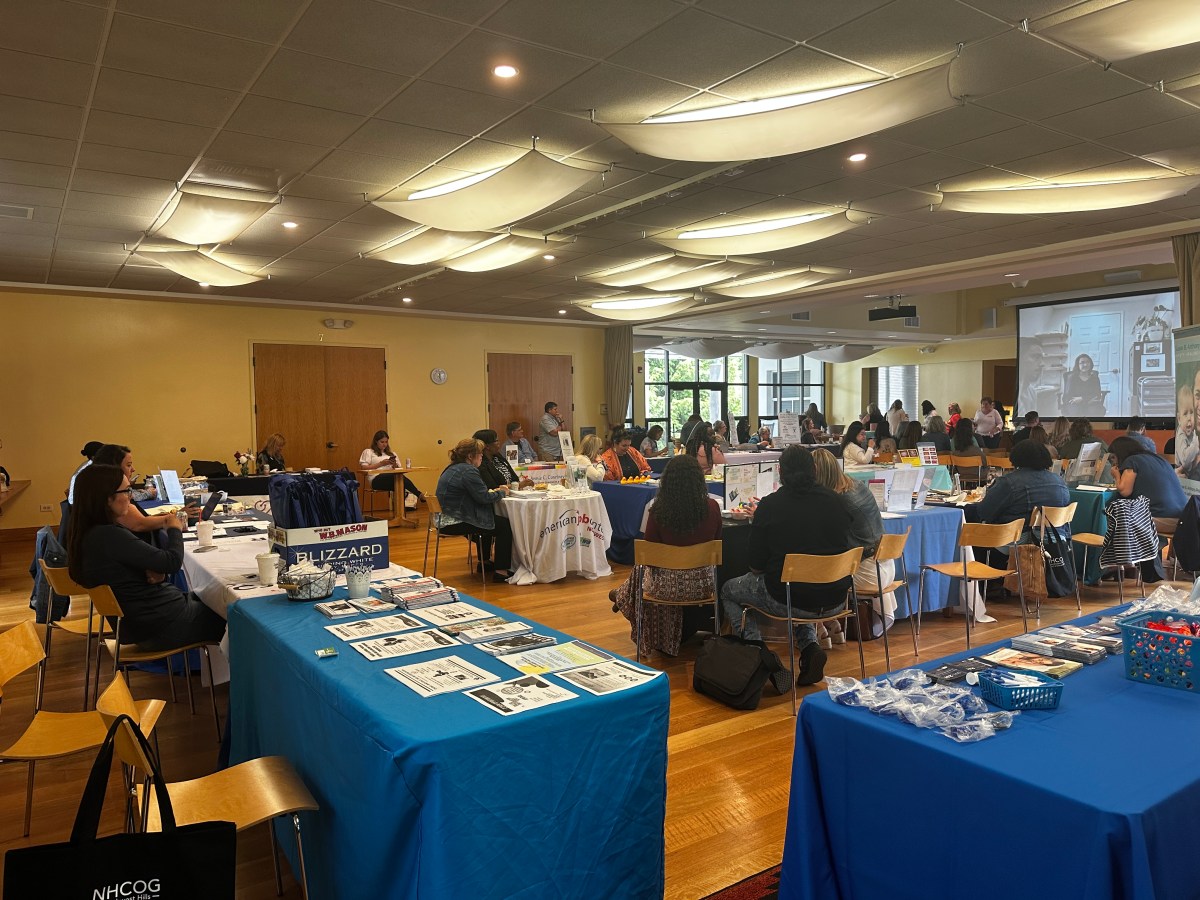 A roomful of people seated at tables with blue and white tablecloths. Attendees look towards a screen in the front of the room.