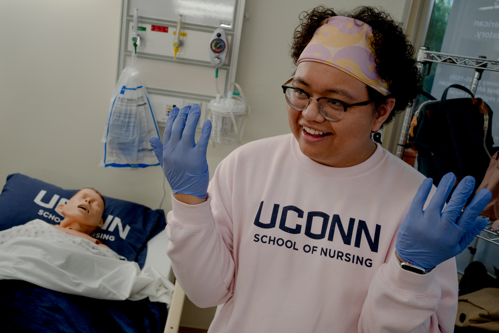 Zaheer Turtem stands with his hands elevated near his shoulders. He is wearing a UConn t-shirt, glasses, a headband and blue plastic gloves.