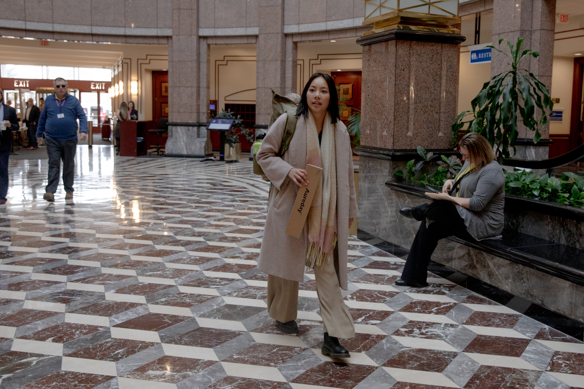 Kimberly Nguyen is walking in the lobby of the Legislative Office Building. She is wearing a a long coat, long pants, boots, and a scarf. She is holding a small cardboard box.