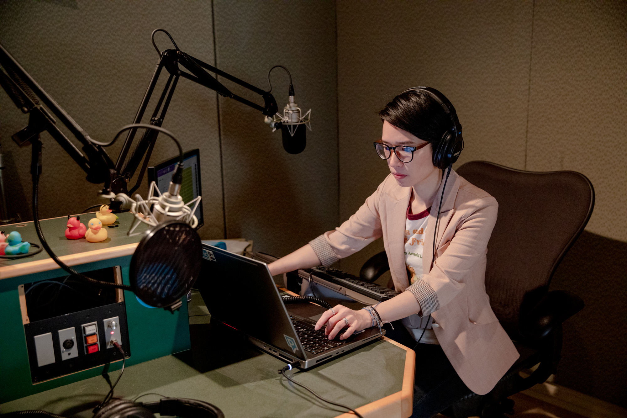 Catherine Shen works at a laptop. She is wearing a blazer, glasses, and a pair of headphones. She is seated in front of a microphone.