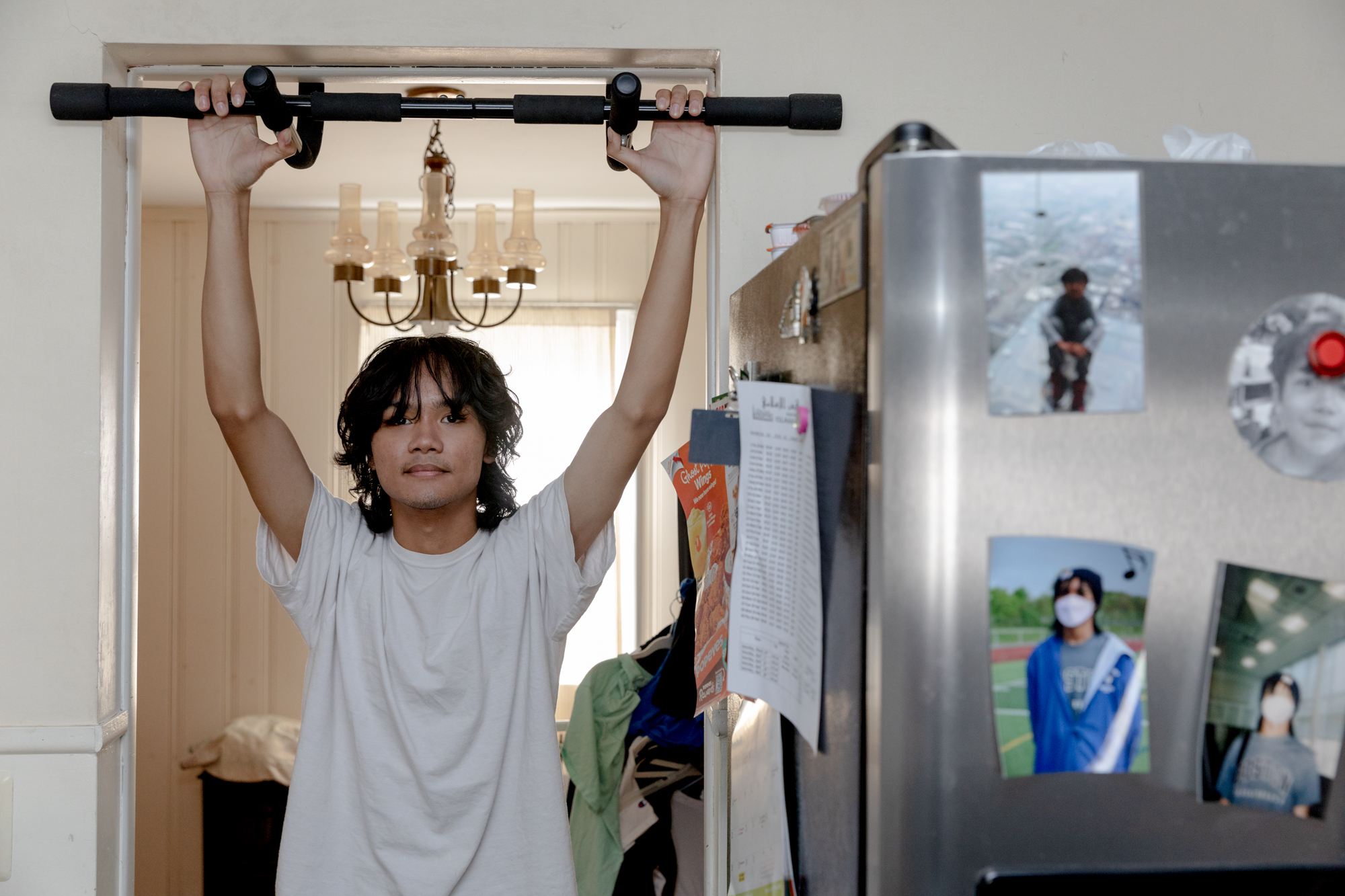 Adam Putra stands in a doorway with his hands extended upwards, holding onto a pull-up bar. He is wearing a white t-shirt.